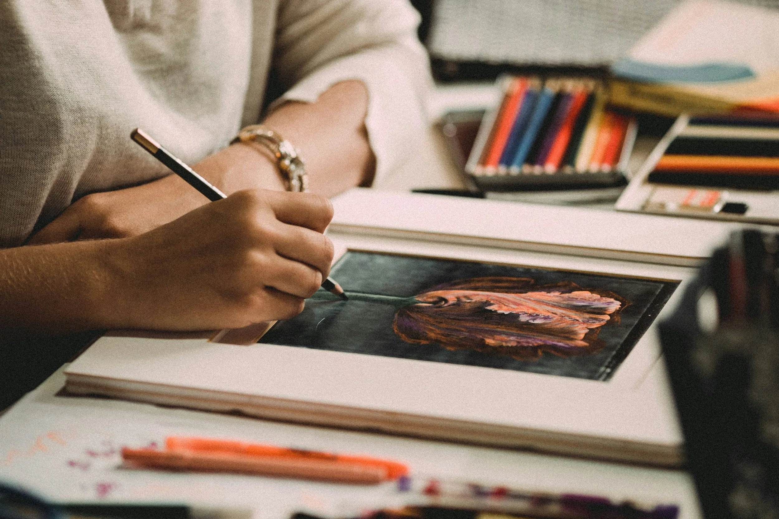A person drawing or painting a colorful, textured piece of artwork on black paper, with art supplies and colorful markers or pastels nearby on a desk.
