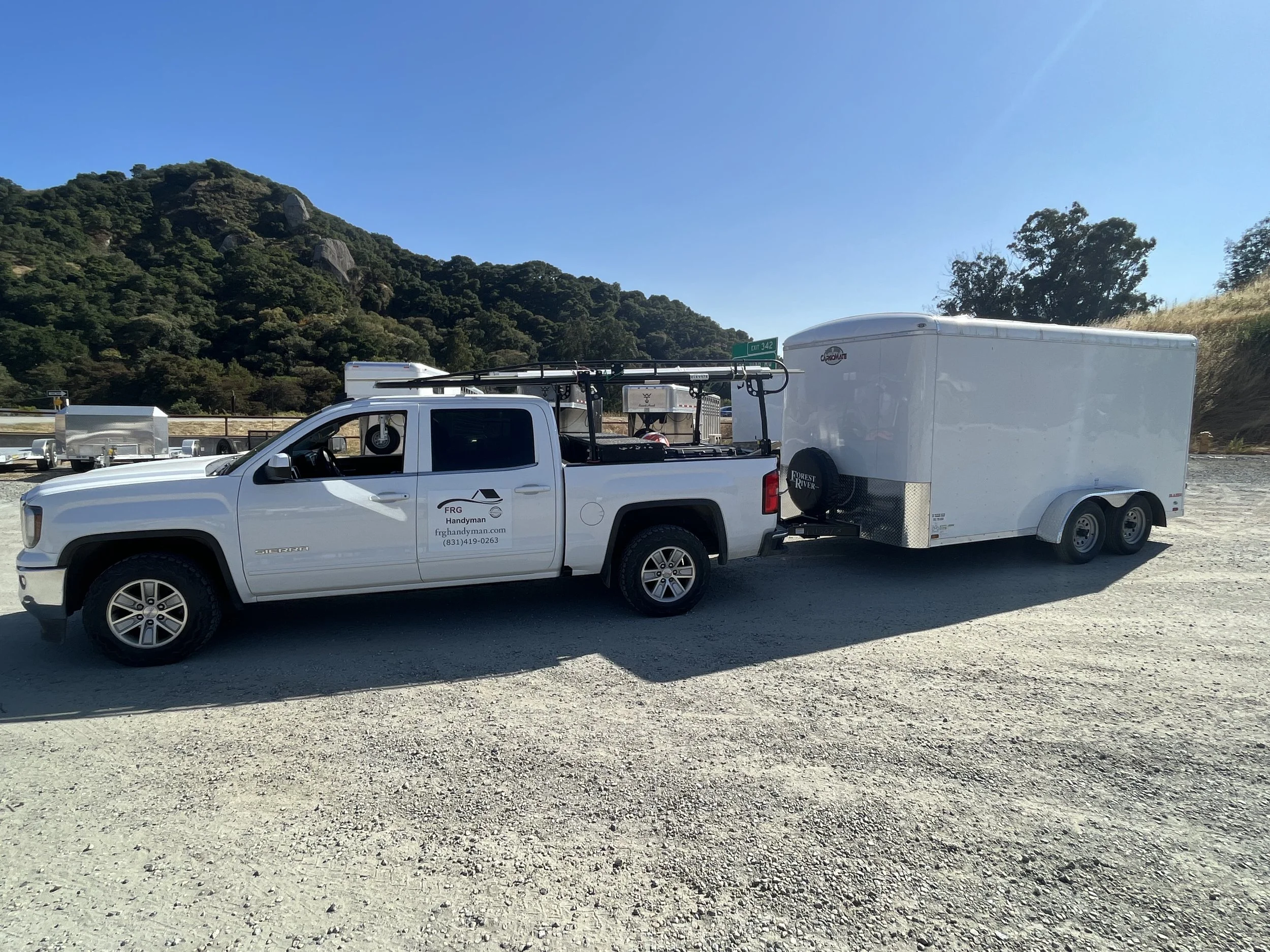 White pickup truck with a logo and contact information on the door, towing a large enclosed trailer, parked on a gravel lot with hills and trees in the background.