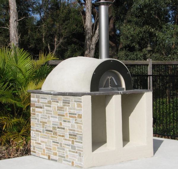 Outdoor pizza oven with a brick facade, chimney, and open oven space, situated on a concrete patio with greenery in the background.