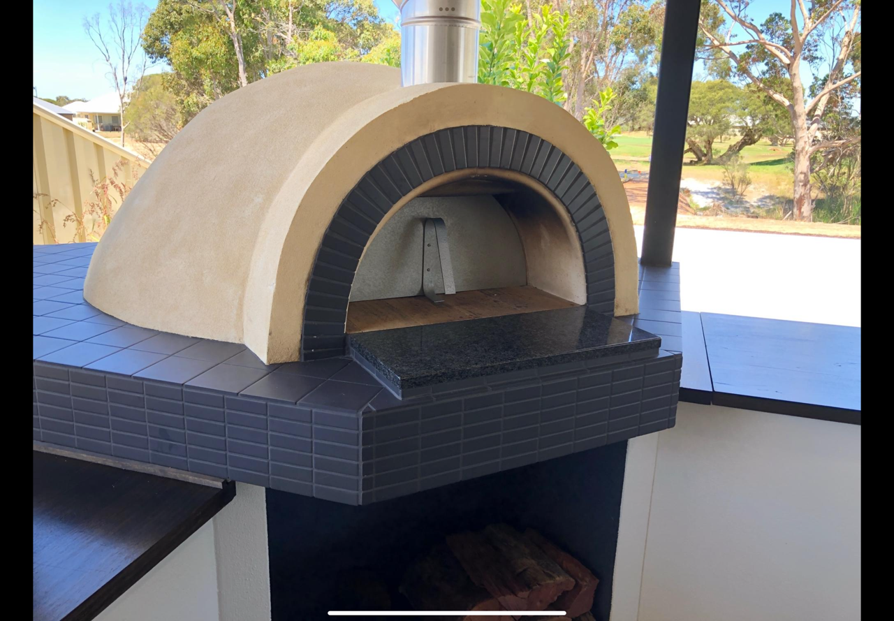 Outdoor pizza oven with a beige dome, black arch detail, and a stone countertop, set against a scenic background with trees and water.