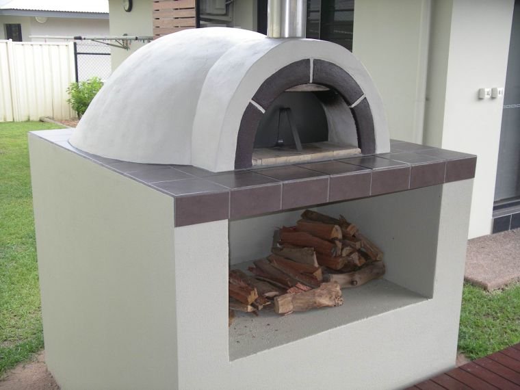 Outdoor pizza oven with firewood stored underneath in a built-in compartment, located in a backyard with a grassy lawn.