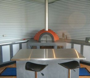 A metallic table surrounded by three black stools in a room with metallic walls and a curved orange oven in the background.