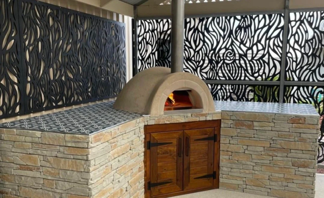 Outdoor pizza oven with stone base, wooden cabinet doors, black decorative metal panels, and tiled countertops.