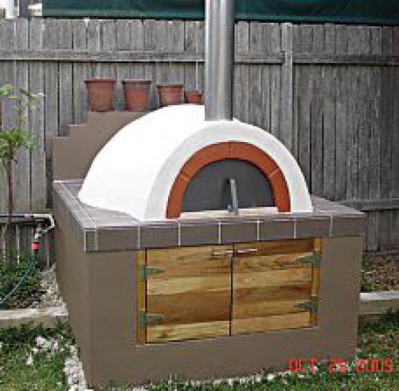 Outdoor pizza oven with a dome top, clay brick accents, and a wooden door on the front, situated in a backyard with a wooden fence and potted plants.