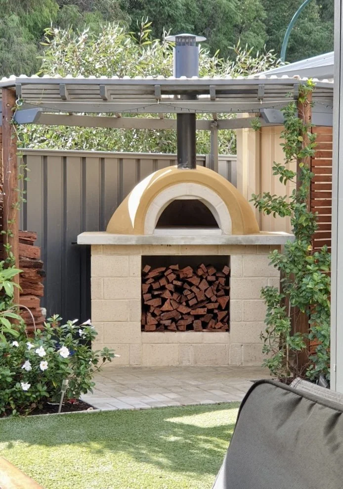 Outdoor backyard pizza oven with wood storage, surrounded by plants and a patio area.