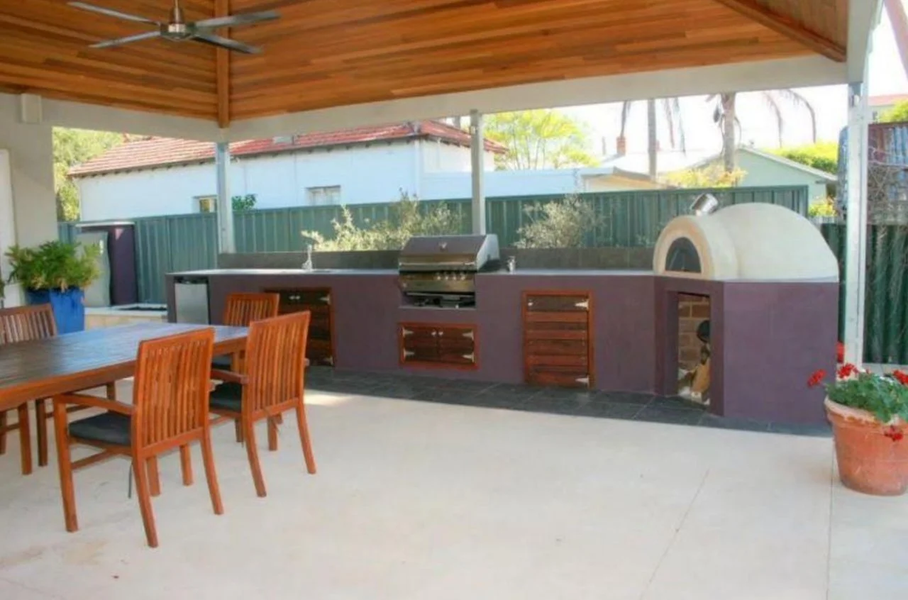 Outdoor patio with a wooden dining table, six chairs, a stone pizza oven, a grill, and potted plants, under a wooden ceiling with a ceiling fan.