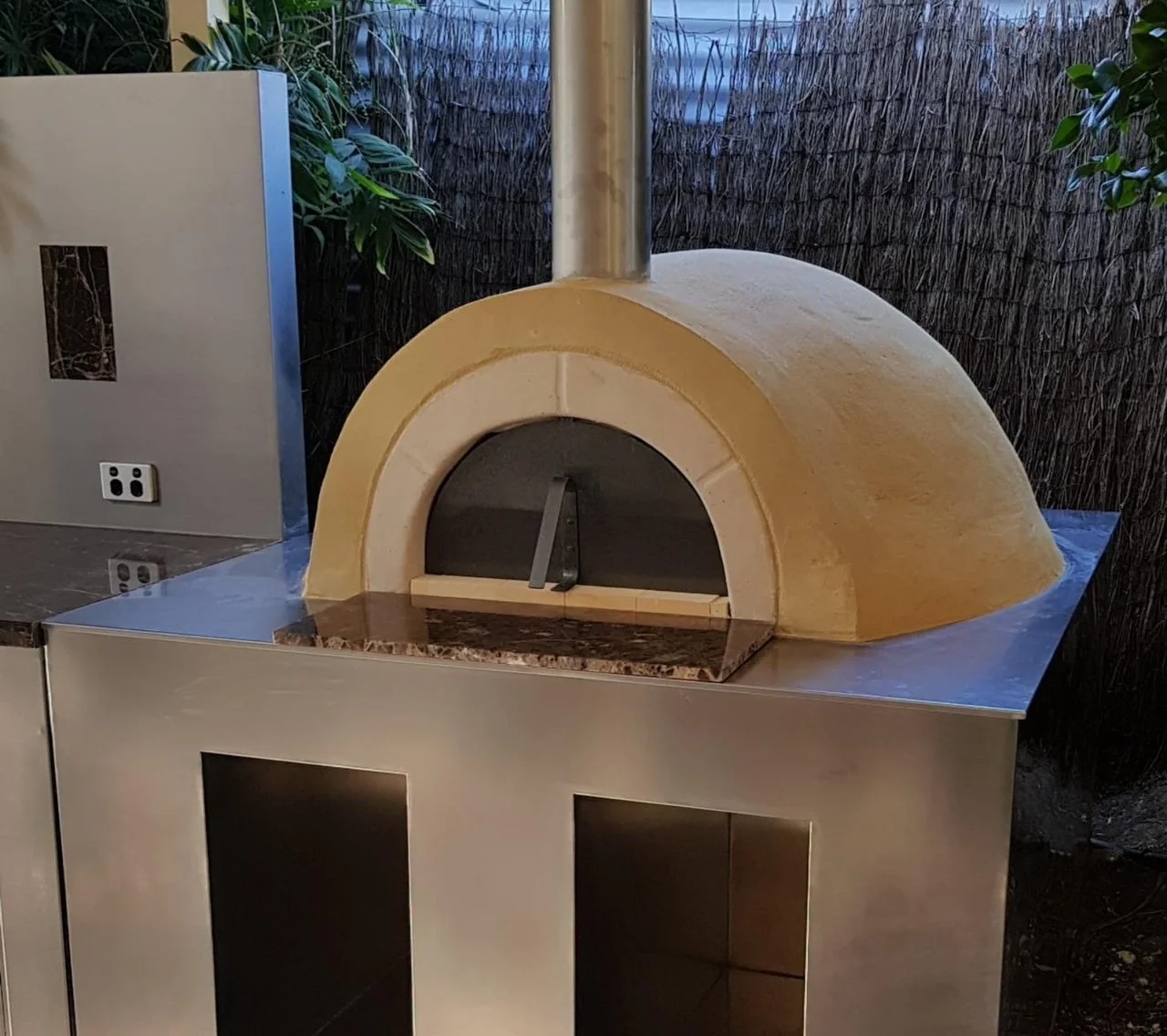 A wood-fired pizza oven with a dome shape, a small chimney, and a temperature gauge on a stainless steel countertop outdoors, surrounded by plants and fencing.