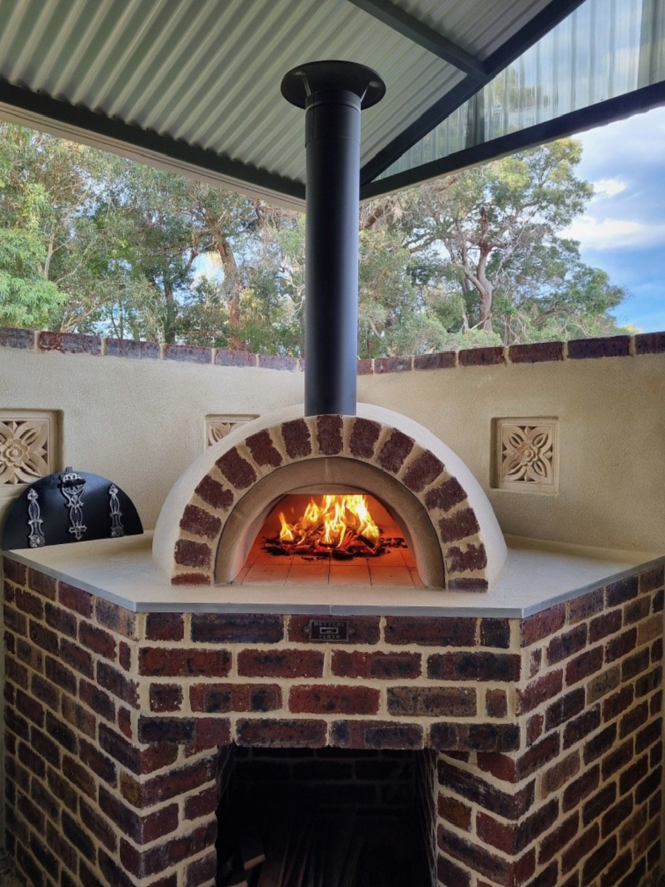Outdoor brick pizza oven with flames inside, situated on a counter with a decorative grill on the left side, under a roof with trees and sky in the background.