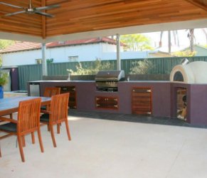 Outdoor kitchen with barbecue grill, wooden cabinets, dining table, and chairs under a covered patio.