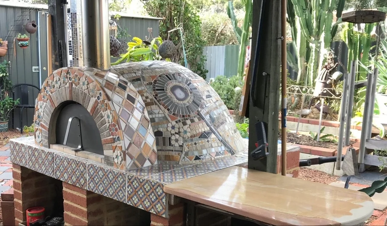 A mosaic pizza oven on a brick stand outdoors with gardening tools and plants around.