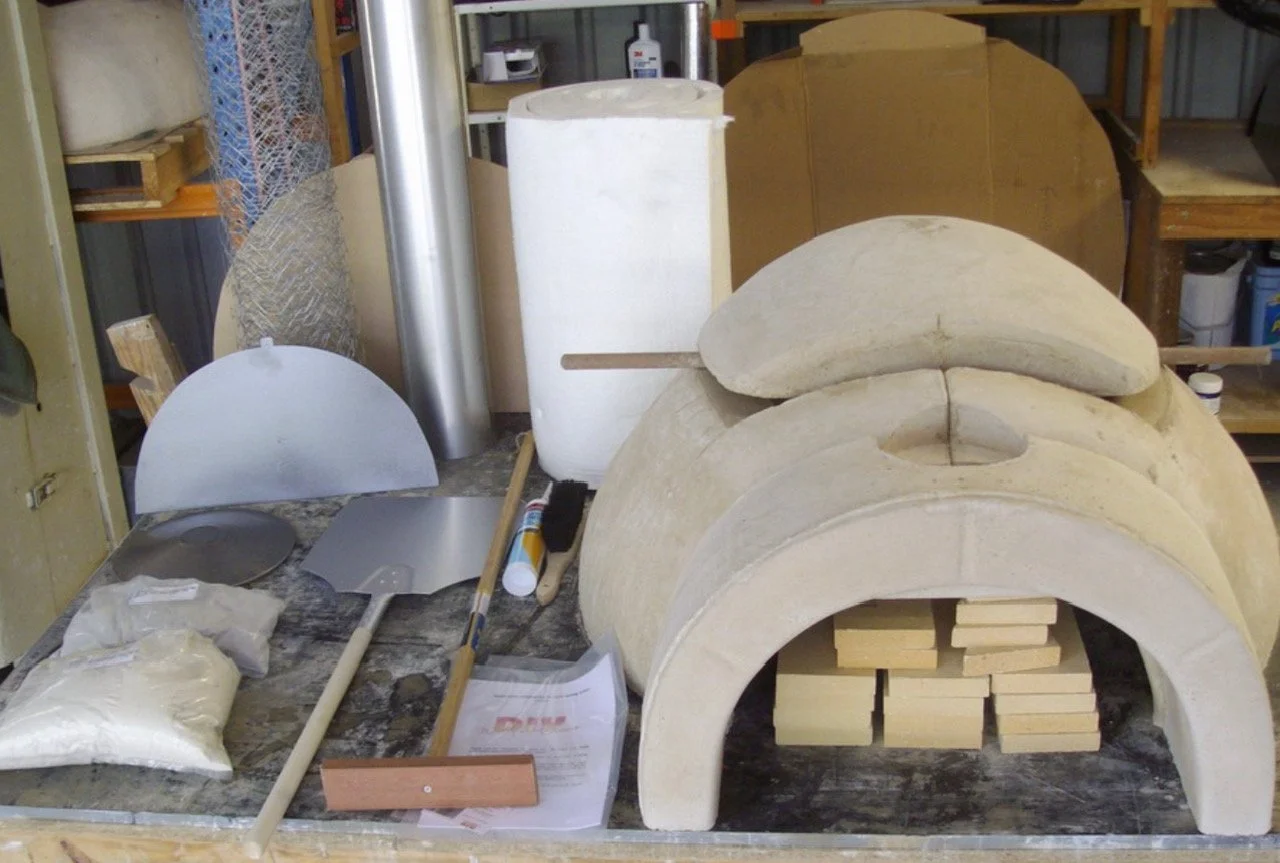 Workshop setup with a small clay pizza oven, construction tools, a bag of clay, and wooden firebricks.