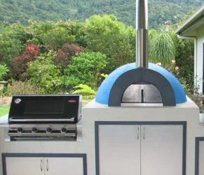 Outdoor kitchen with a built-in pizza oven and a gas grill on a white countertop, surrounded by greenery and a house in the background.