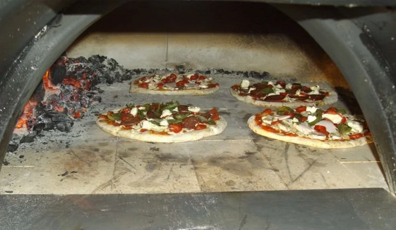 Four small pizzas baking in a wood-fired oven with glowing embers.