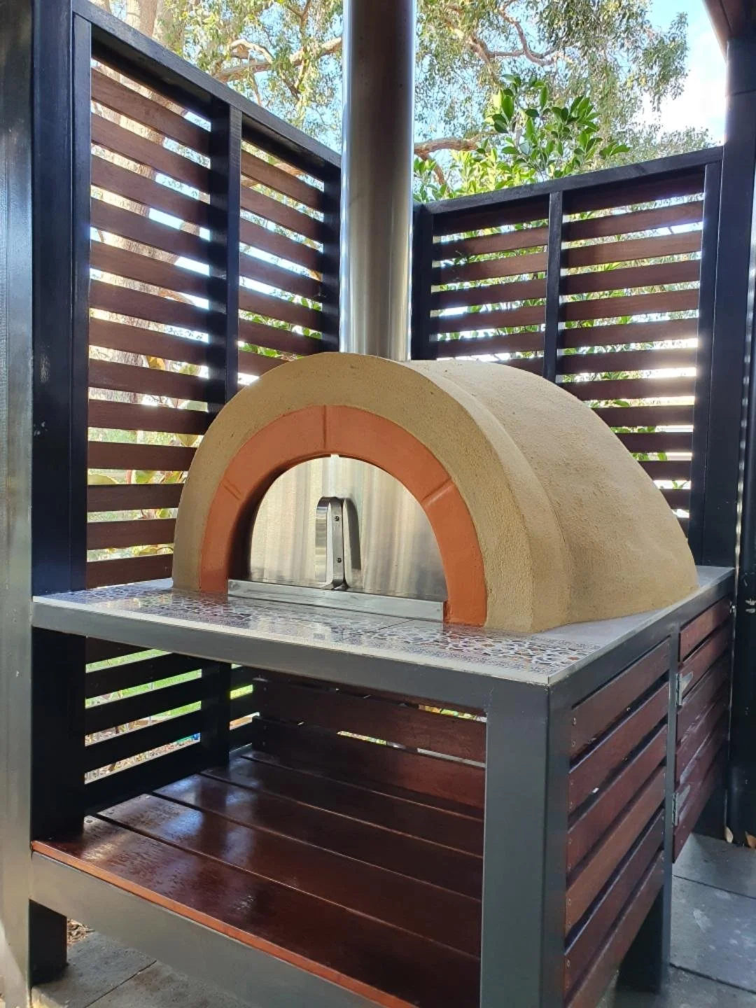 Outdoor pizza oven with a curved, brick-lined opening, set on a tiled surface, surrounded by wooden slatted fencing and greenery in background.