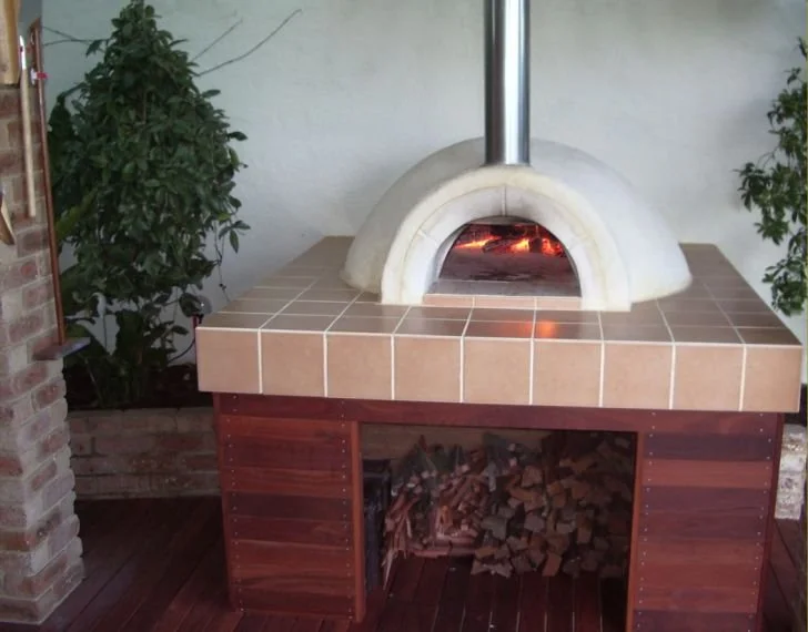 Outdoor pizza oven with a fire inside, built on a tiled countertop with wooden storage underneath, surrounded by potted plants.