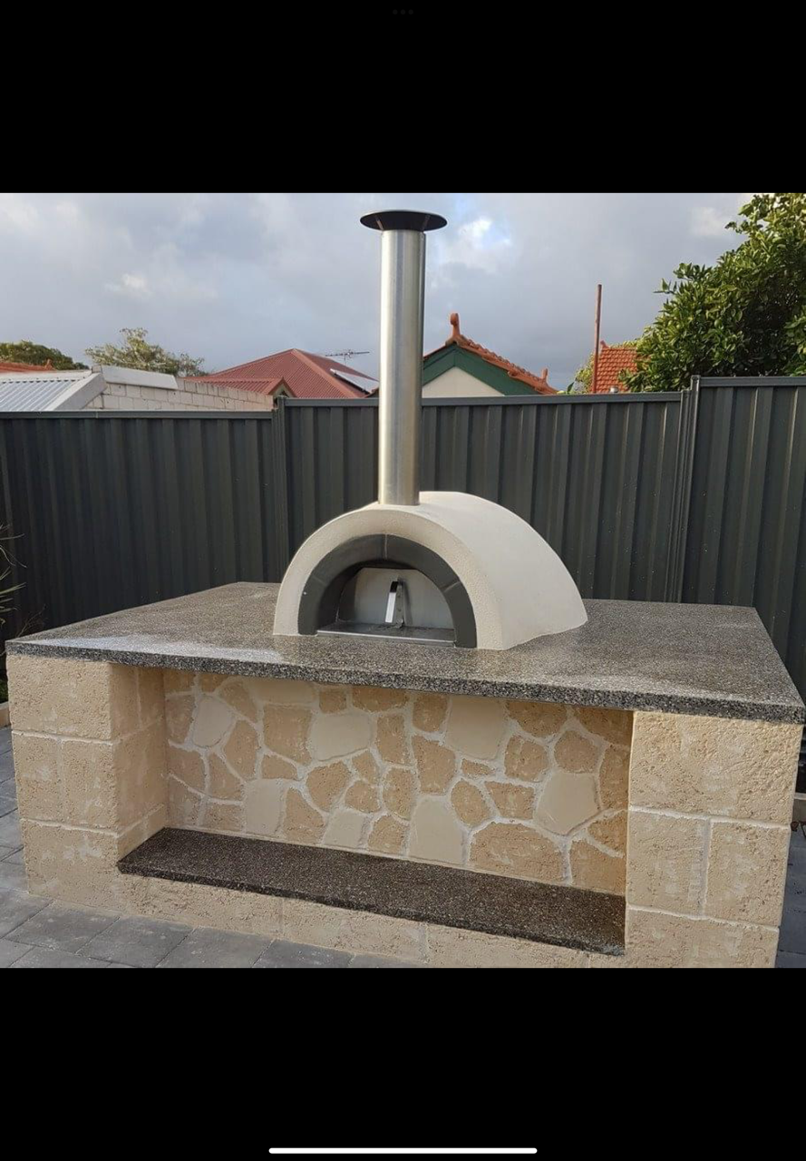 Outdoor pizza oven with a stone and concrete base, a domed oven with a metal chimney, set against a backyard fence with houses in the background.
