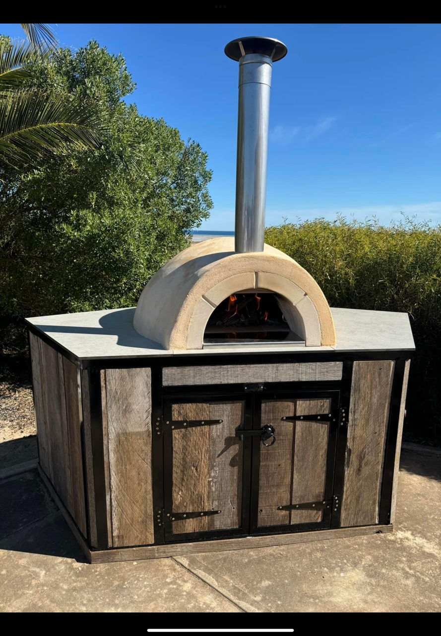Outdoor pizza oven with a chimney, set on a wooden counter, on a patio with greenery and blue sky in the background.