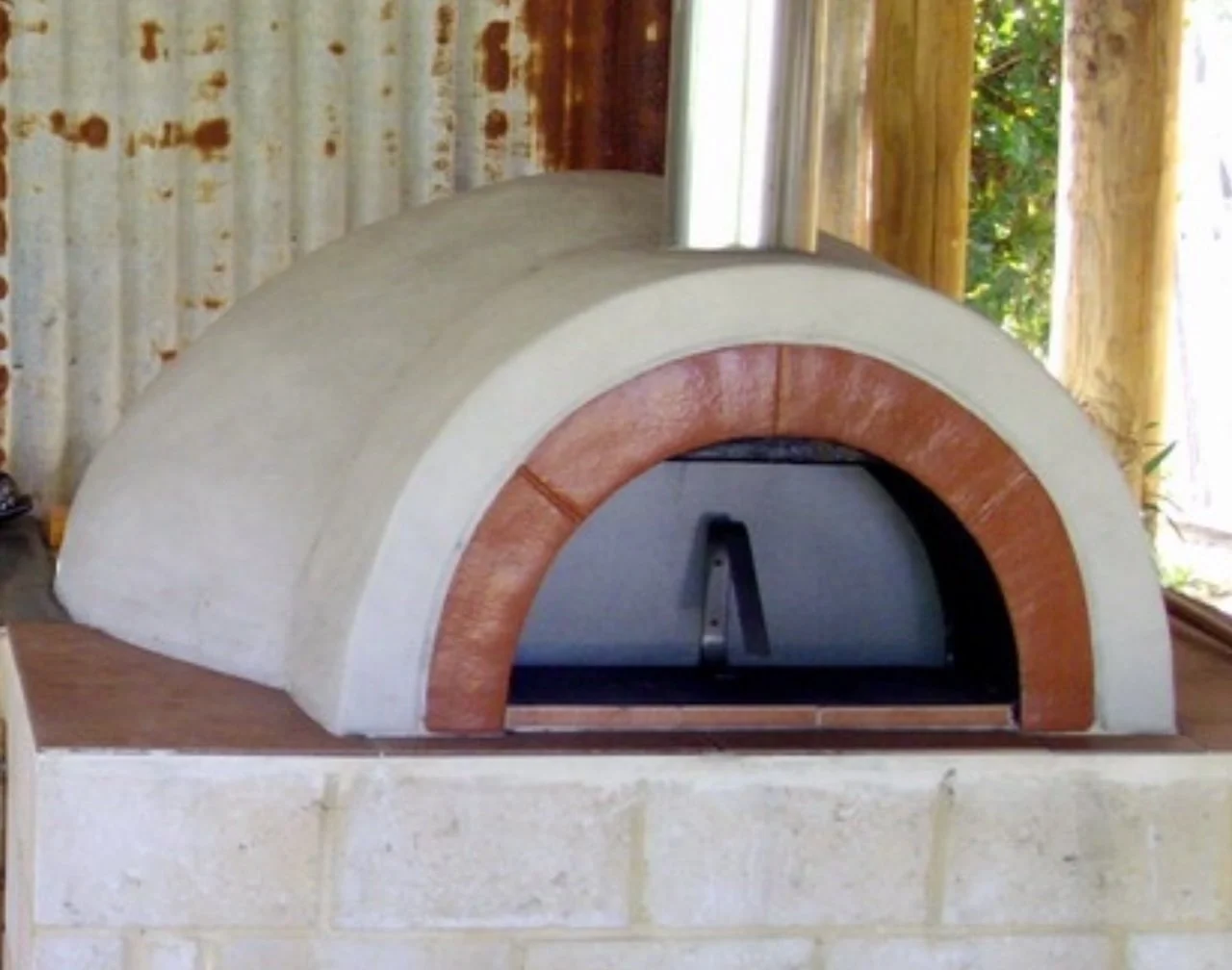 A wood-fired pizza oven with a dome shape, surrounded by bricks, in an indoor setting with wooden and metal elements and natural greenery visible through windows.
