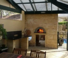 Outdoor patio with fireplace, chairs, and a wooden table. Covered by a roof with a screened area, with greenery outside.