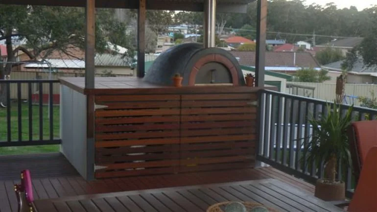 Outdoor deck with a tiled outdoor pizza oven, wooden railing, and potted plant, overlooking a suburban neighborhood with trees and houses in the background.