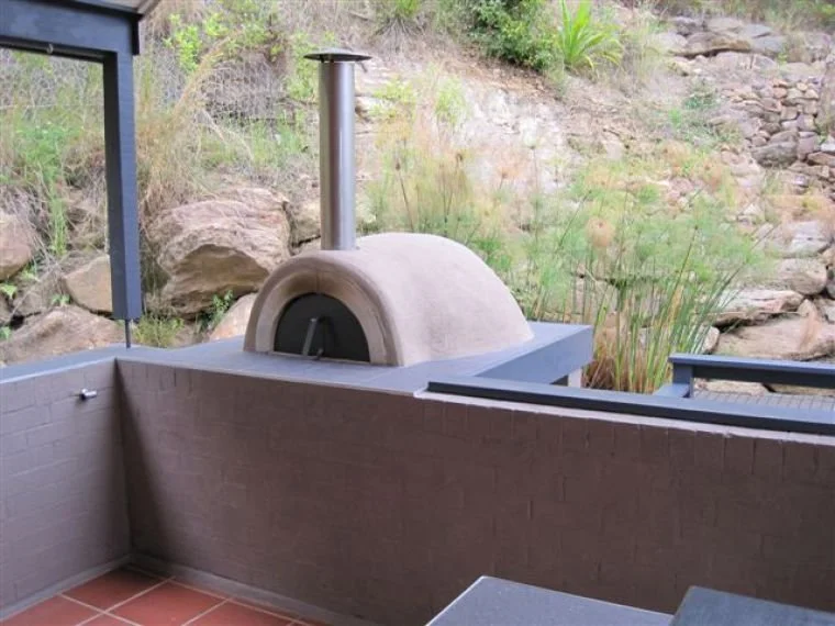 Outdoor pizza oven with a chimney on a patio with brick wall, plants, and rocky hillside in the background.