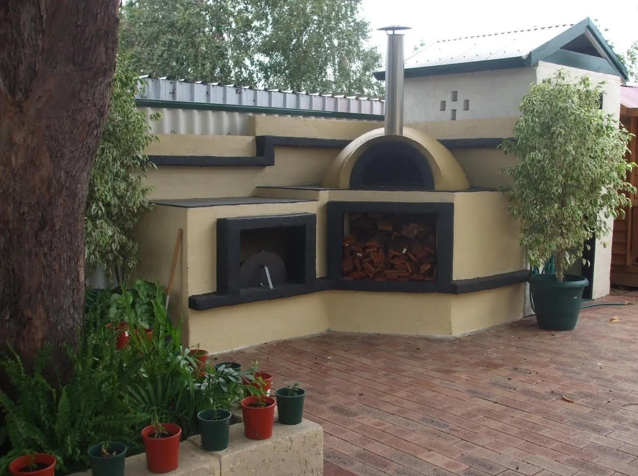 Outdoor pizza oven with a chimney, built into a stucco structure with wood storage, on a brick patio, with potted plants and trees nearby.