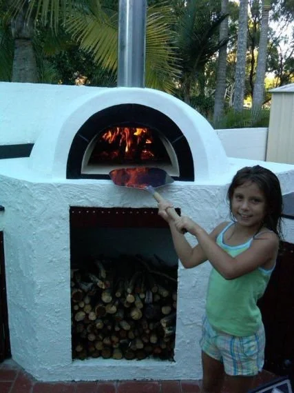 Young girl using a pizza peel to place pizza into outdoor stone pizza oven with a wood fire inside and a stack of firewood below, in a backyard with trees.