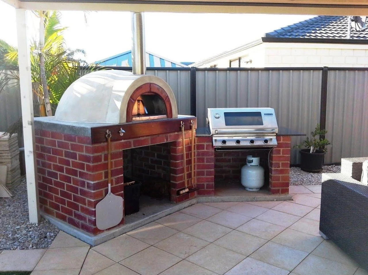 Outdoor kitchen with a brick pizza oven, a stainless steel barbecue grill, and a propane tank underneath the grill, located on a tiled patio with potted plants and a privacy fence.