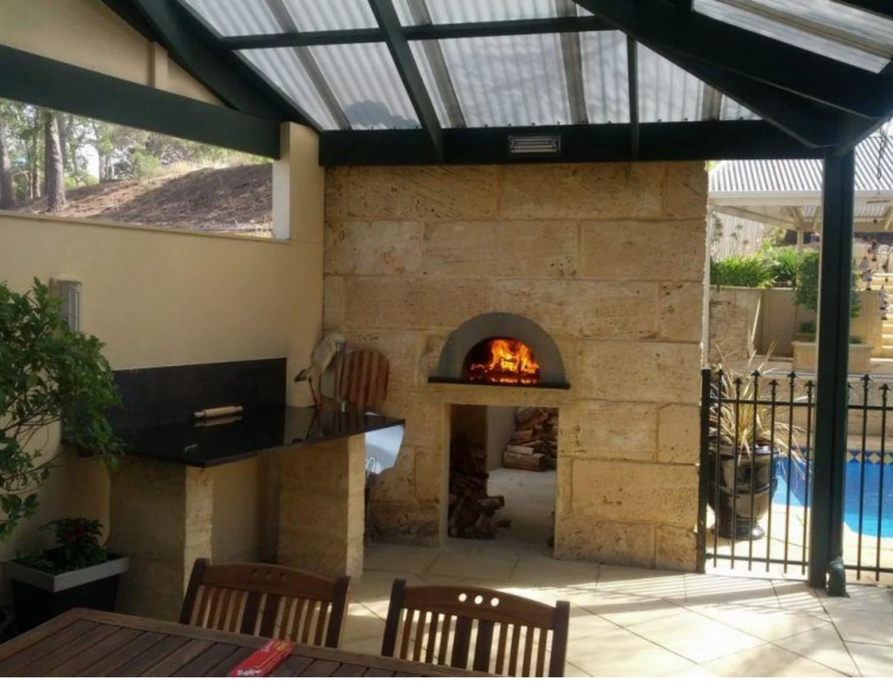 Outdoor patio with stone fireplace, wood fire burning, barbecue grill, glass roof, wooden table and chairs.