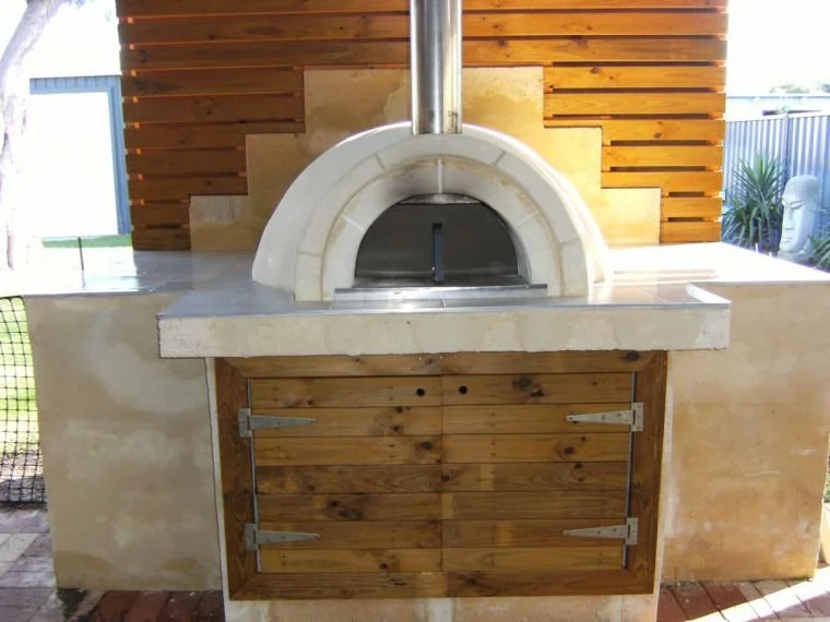 An outdoor pizza oven with a stone dome and a metal chimney, mounted on a concrete counter with a wooden door beneath, and a wooden wall in the background.