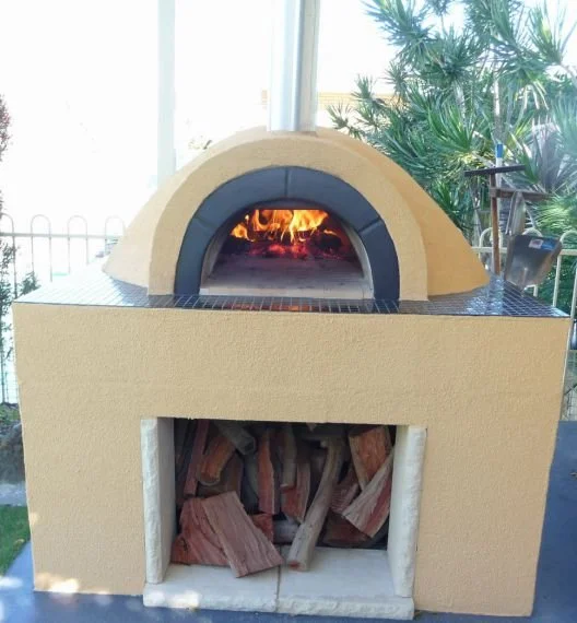 A backyard pizza oven with a fire inside, built into a yellow stucco structure with firewood stored beneath, and surrounded by plants and outdoor furniture.