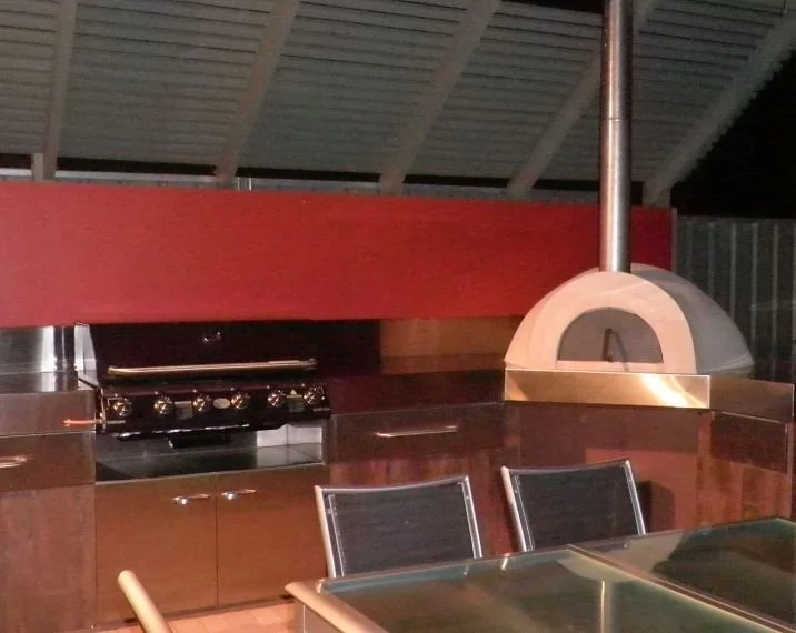 An outdoor pizza oven with a wood-fired design next to a stainless steel grill, situated on a patio with metal chairs and a glass table.
