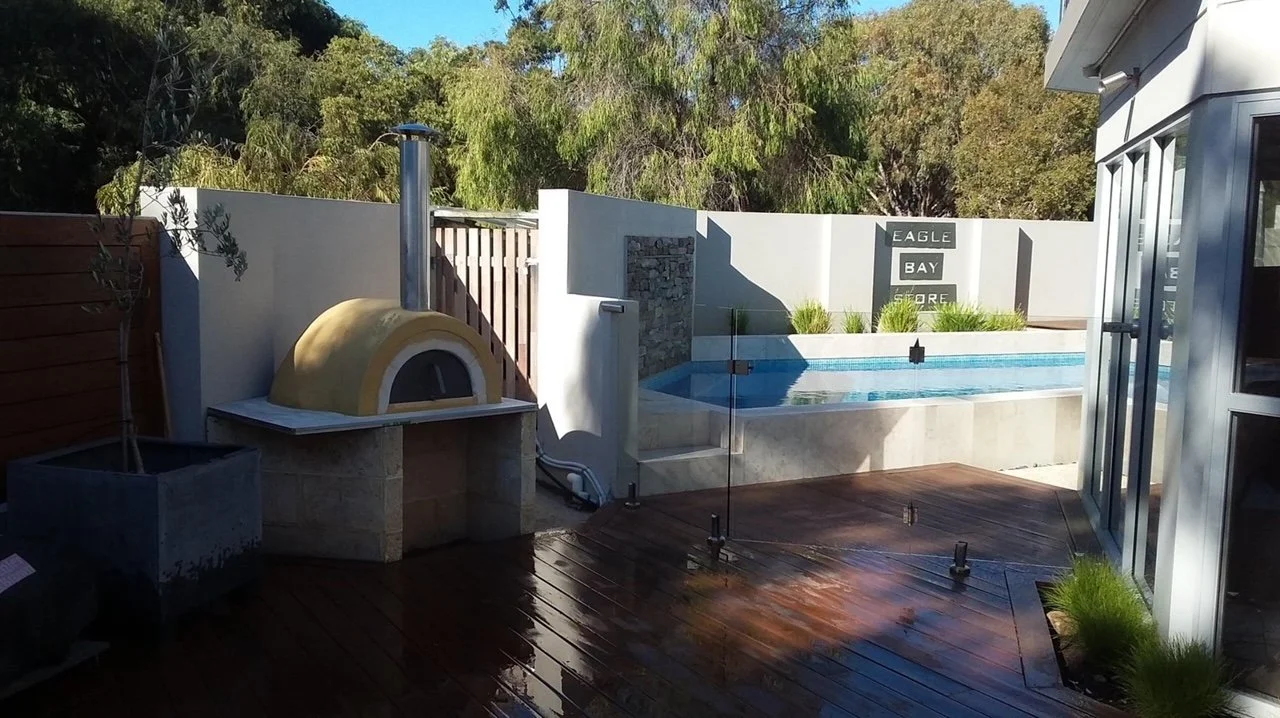 A backyard with a modern pool, a small pizza oven, and a glass fence. The area has a wooden deck and is surrounded by trees and greenery.