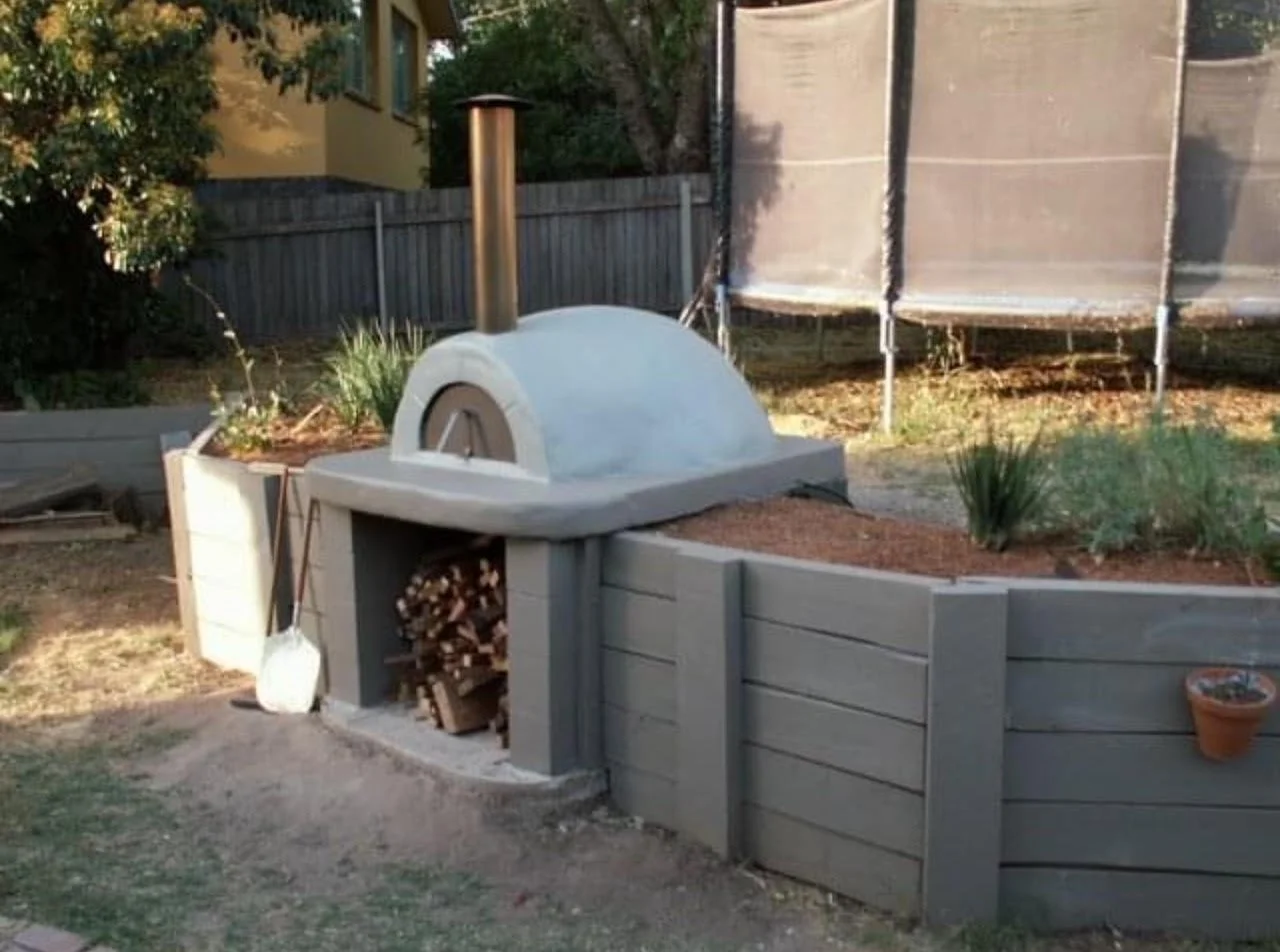 A backyard with a wood-fired pizza oven in a raised planter bed, surrounded by gardening plants, with a wood pile underneath the oven and a shovel leaning against it.