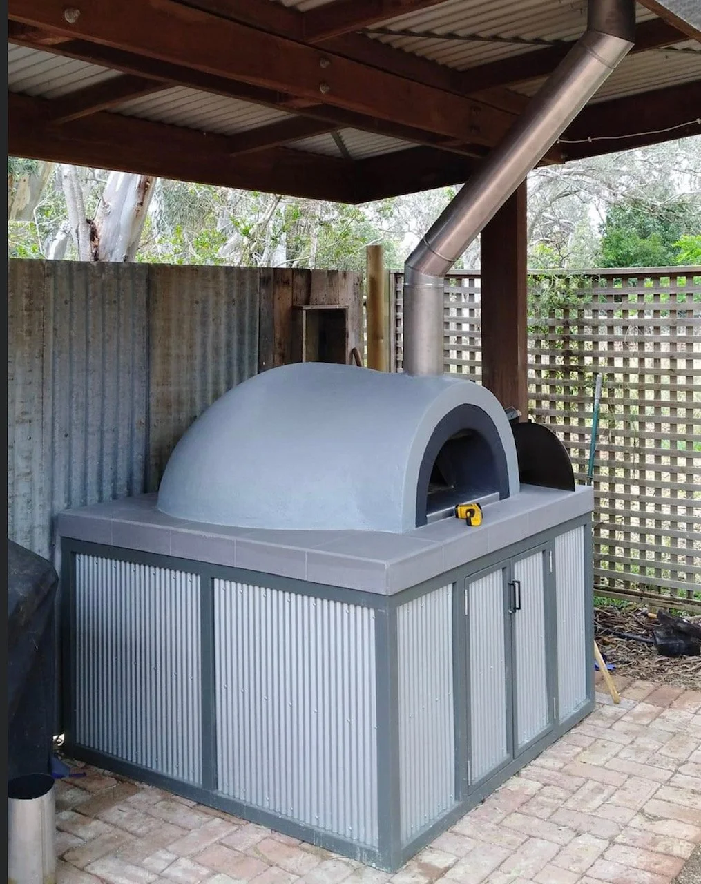 Outdoor pizza oven with a metal chimney and a storage cabinet underneath, situated on a brick patio with a wooden fence and trees in the background.