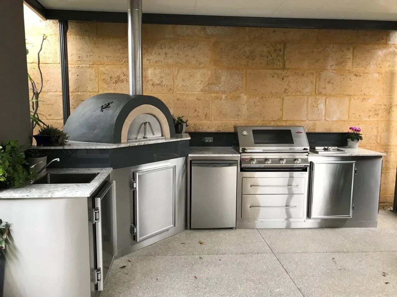 Outdoor kitchen with pizza oven, grill, and side cabinets, decorated with potted plants, against a brick wall.