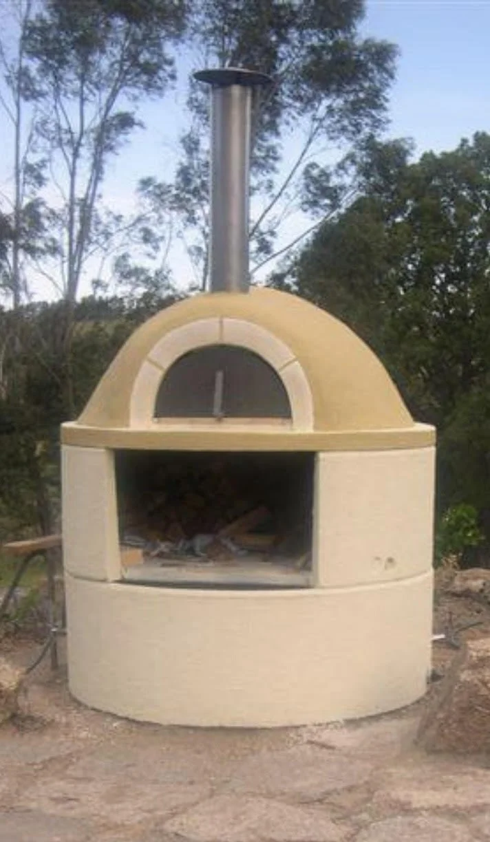 An outdoor stone or brick pizza oven with a metal chimney, set against trees and a blue sky.