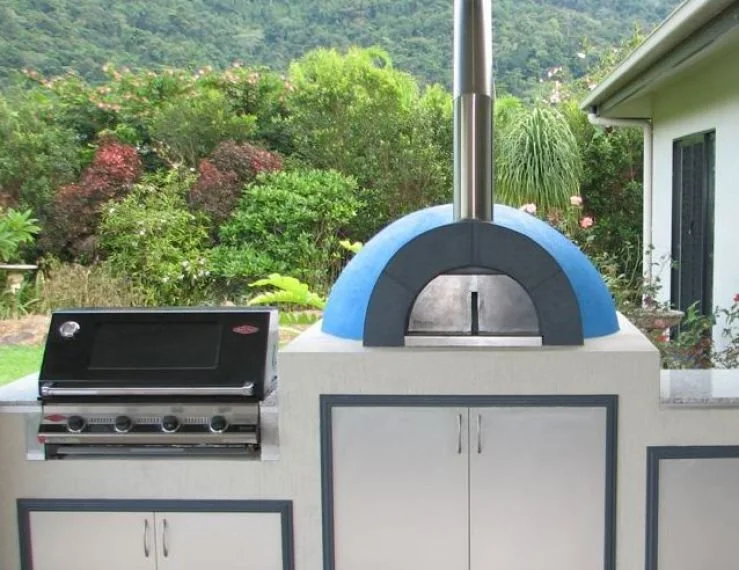 Outdoor pizza oven with a dome-shaped top, a built-in pizza peel, next to a barbecue grill, set against a lush garden backdrop.