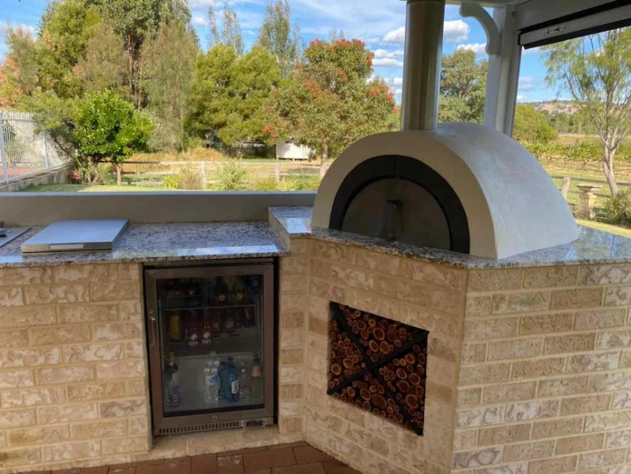 Outdoor kitchen with brick counter, a pizza oven, a mini fridge, and firewood storage, with a scenic view of trees and a partly cloudy sky in the background.