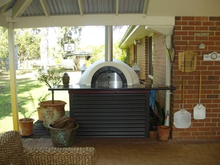 An outdoor brick and concrete pizza oven with a black metal door, located under a porch with a metal roof. To the left of the oven, there are potted plants and a small sculpture, with a wicker chair in the foreground. To the right, hanging on a brick