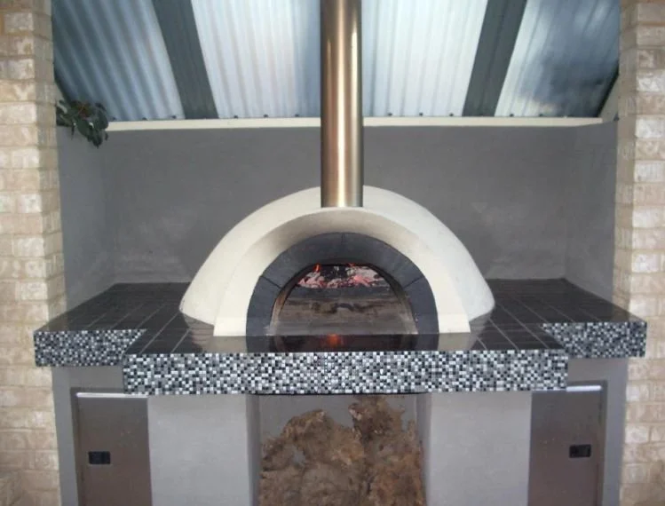 Outdoor pizza oven with a dome shape, topped with black and white mosaic tiles, situated on a tile countertop with a metal chimney pipe.