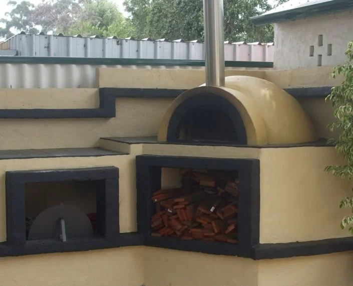 Outdoor wood-fired pizza oven with a chimney and storage compartments, one filled with firewood, on a yellow stucco wall.