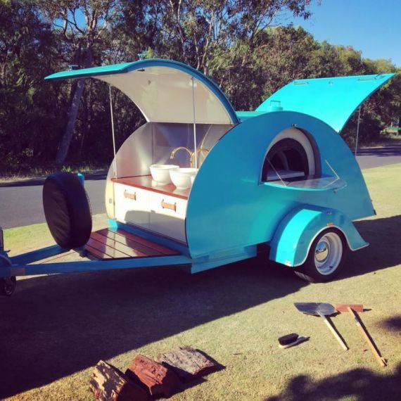 A small, blue, teardrop-shaped trailer with an open side revealing an interior with a table and bowls, parked on a grassy area with trees in the background.