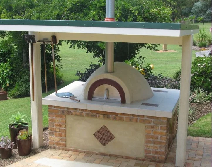 Outdoor brick pizza oven with a white countertop, under a small roof, surrounded by a garden with plants and grass.
