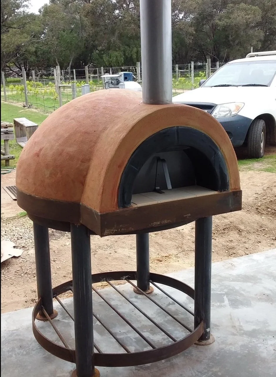 A small outdoor pizza oven with a round dome-shaped terracotta exterior, a black opening, and a metal chimney, set on a metal stand with a circular shelf, in a backyard with cars and a vineyard in the background.