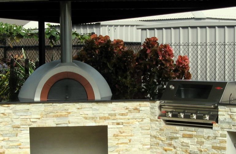 Outdoor pizza oven, mounted on a stone countertop, with a garden and plants in the background, and a barbecue grill to the right.