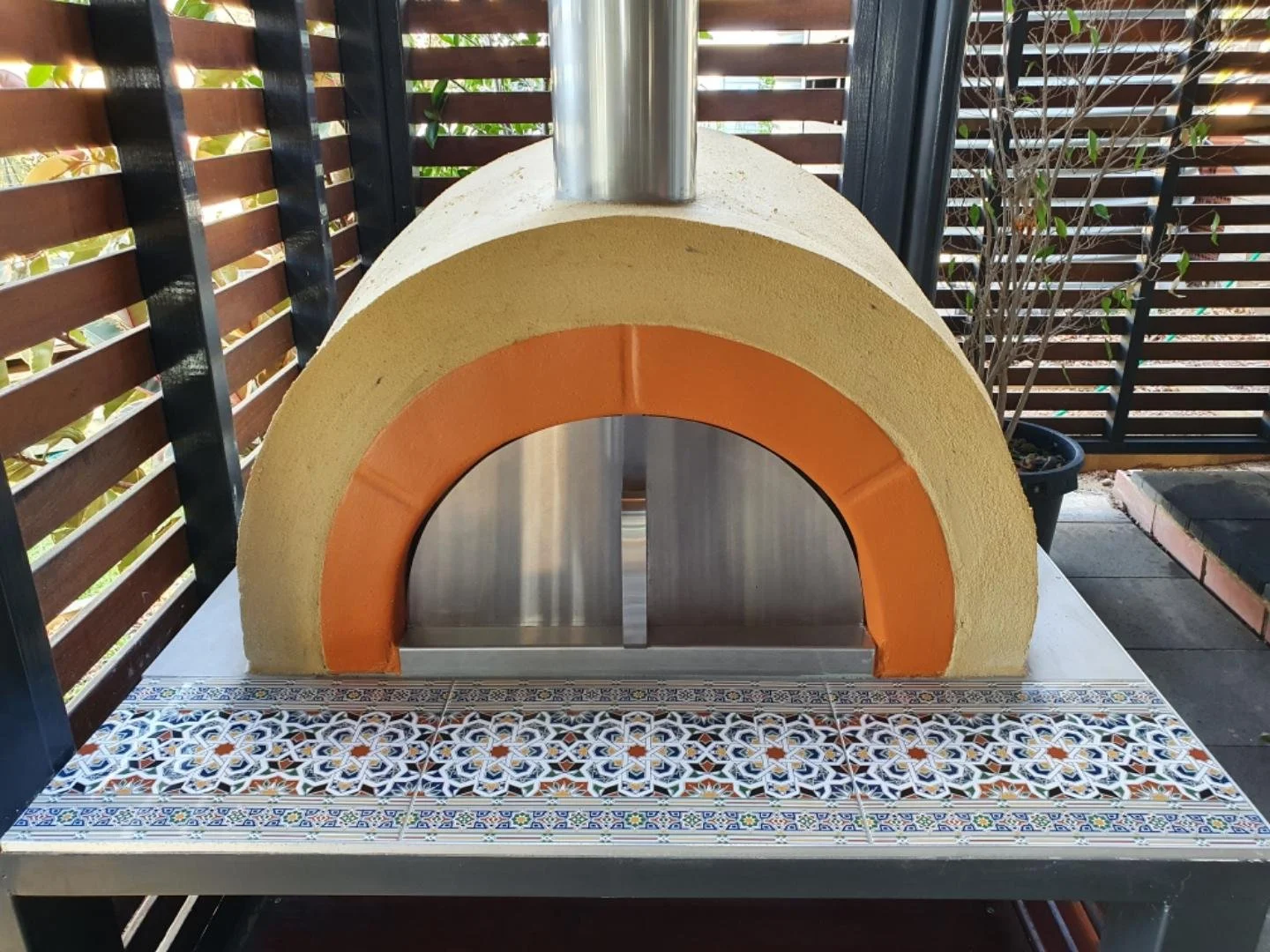 A modern outdoor pizza oven with a yellow exterior, orange accents around the opening, and a metallic chimney on top, placed on a decorative tiled table on a patio.