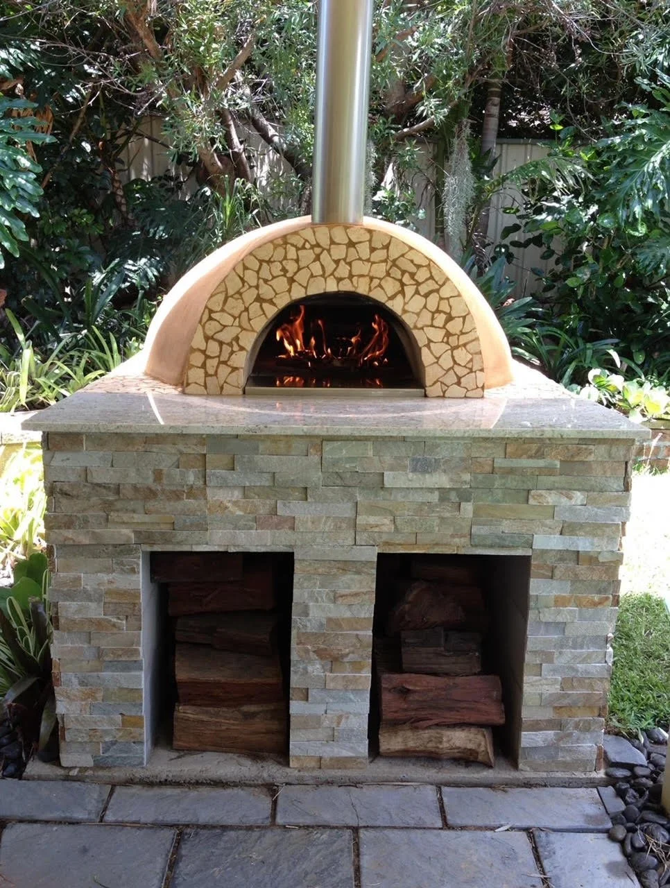 Outdoor stone pizza oven with a fire inside, surrounded by lush green plants and trees.