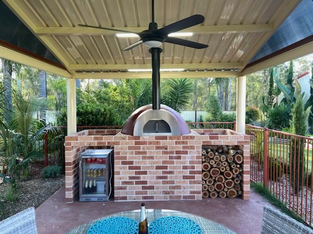 Outdoor brick pizza oven with a black ceiling fan above it, under a covered patio with a metal roof. To the left, there is a small refrigerator, and on the right, there are neatly stacked logs of firewood. The setting is surrounded by lush greenery.
