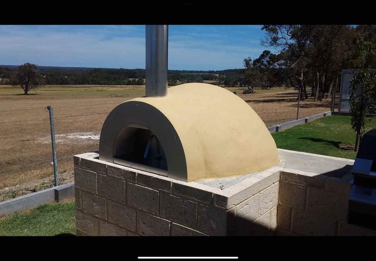 A pizza oven with a brick base and a stone dome roof, located outdoors on a concrete slab with a rural landscape in the background.